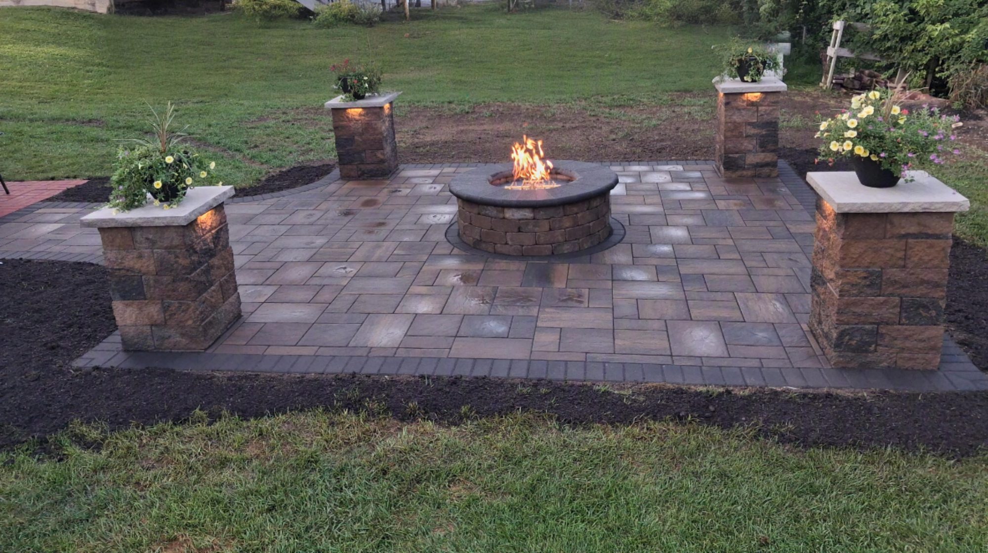 A paver patio by M.S.S Landscaping with a round gas fire pit and four lit stone pillars